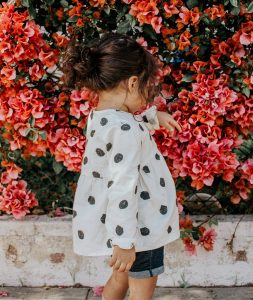 girl in black and white polka-dot blouse and blue shorts standing beside red petaled flowers during daytime